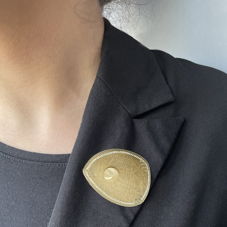 Broche en corne dorée, ornée d’une délicate frise composée de deux rangées de petits points blancs peints à la main. Une pastille de nacre ivoire apporte lumière et éclat. La broche est portée ici sur une veste noire.