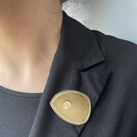 Broche en corne dorée, ornée d’une délicate frise composée de deux rangées de petits points blancs peints à la main. Une pastille de nacre ivoire apporte lumière et éclat. La broche est portée ici sur une veste noire.