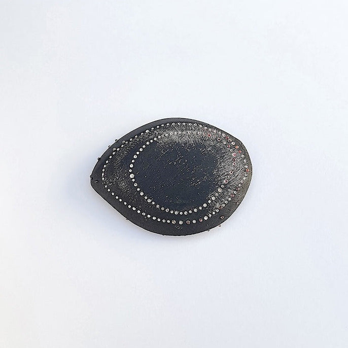 Cette broche en ébène sculpté, patiné et texturé révèle deux formes ovales dessinées par des points blancs et rosés peints à la main, rappelant l'esthétique des dessins aborigènes.