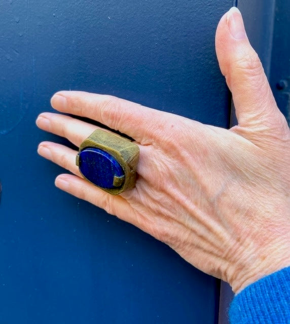 Bague sculpturale rectangulaire, en bronze au centre un lapis-lazuli taillé ovale est serti par deux griffes de chaque côté. Bague portée, décor bleu. 