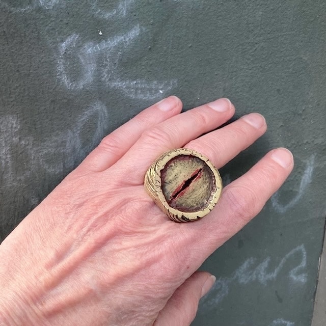 Chevalière sculpturale ronde en bronze , une entaillé au centre dans le métal est soulignée de peinture acrylique rouge.  Les flancs la bague sont griffés de noir. Bijou porté, décor mur vert.