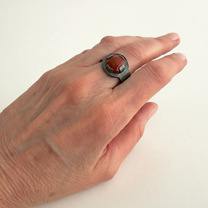 Bague architecturale forme chevalière en argent oxydé avec sertissage contemporain autour d'un jaspe rouge facetté. Détail bijou porté.