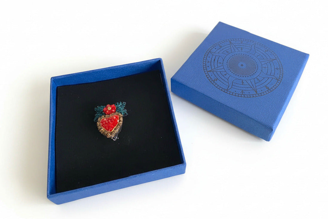 Prête à offrir, La broche Coeur de houx est présentée dans une boîte bleue signée Trovelore, accompagnée de sa notice explicative détaillant le travail de broderie et le savoir-faire artisanal.