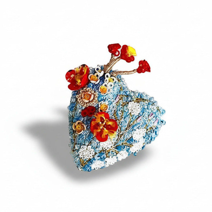 Broche textile contemporain, Heart Blossoms, est un bijou au style floral brodé à la main au fil avec des mini perles de verre et des sequins, dans des tons de bleu pastel. Une branche avec trois fleurs rouges apporte contraste et poésie au bijou.