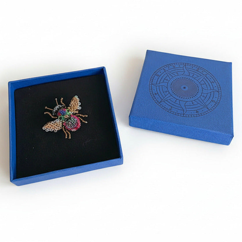 Prête à offrir, La broche Abeille arc en ciel est présentée dans une boîte bleue signée Trovelore, accompagnée de sa notice explicative détaillant le travail de broderie et le savoir-faire artisanal.