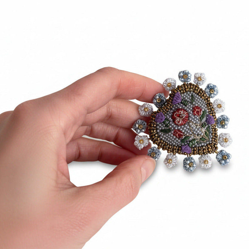 Fabriquée à la main par l'atelier Trovelore, la Broche Coeur Patrimoine est brodée de perles aux fils de couleur dans les tonalité de bleu. De mini fleurs brodées soulignent le bijou présenté par une main.