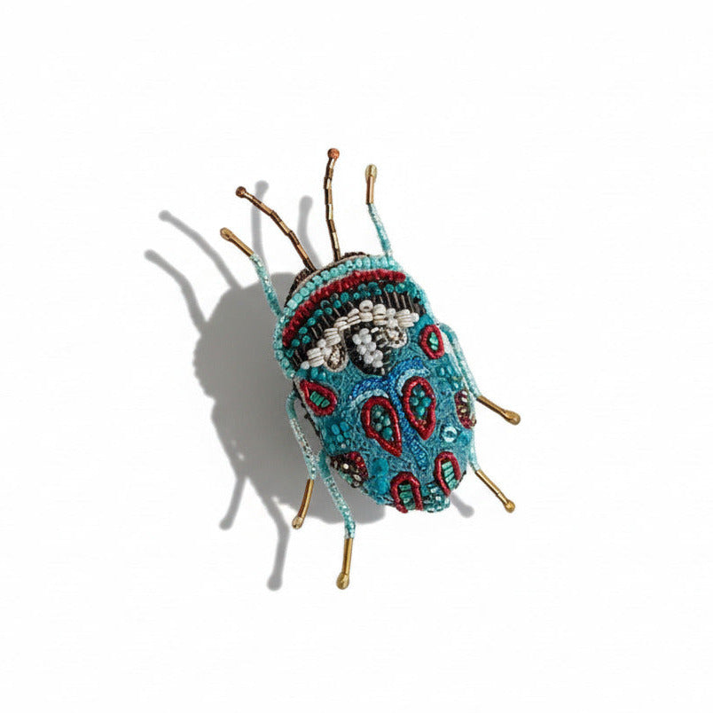 Brodée à la main à l’aiguille, la broche Bug Picasso  traduit en fil, perles et sequins dans des tons de bleu turquoise, blanc et rouge, la richesse graphique de cet insecte iconique.