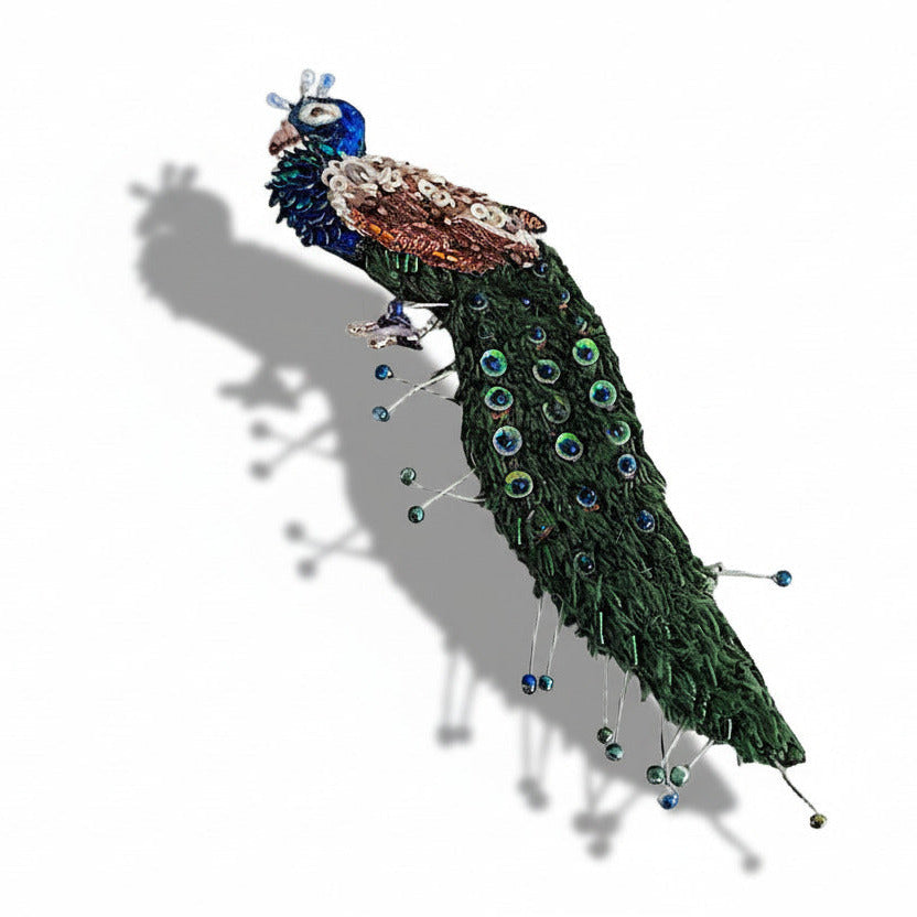 La broche Paon orné de bijoux célèbre la majesté de cet oiseau fascinant. Ce bijou textile est brodé à la main avec des mini perles de verre et des sequins dans des tons de bleu et vert aux reflets irisés.