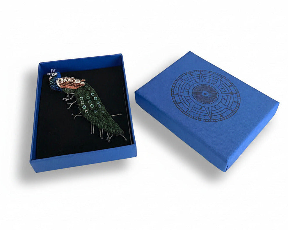 Prête à offrir, La broche Paon est présentée dans une boîte bleue signée Trovelore, accompagnée de sa notice explicative détaillant le travail de broderie et le savoir-faire artisanal.