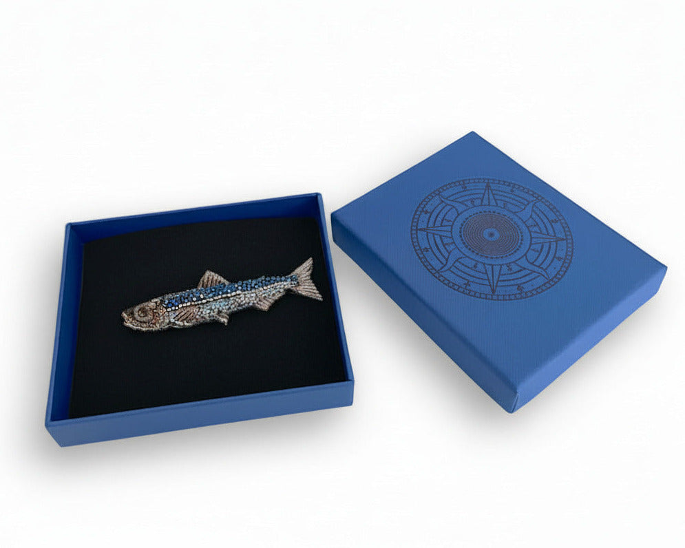 Prête à offrir, La broche Paon est présentée dans une boîte bleue signée Trovelore, accompagnée de sa notice explicative détaillant le travail de broderie et le savoir-faire artisanal.