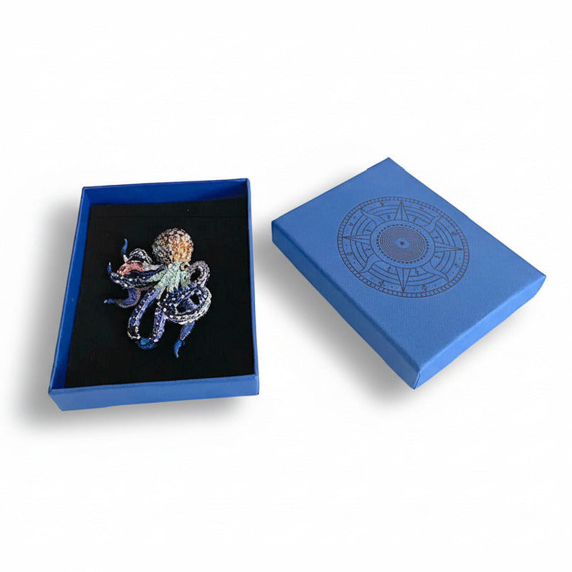 broche sculpturale ornée de mini perles de verre et de sequins, dans des tons de bleu et de brun. Ce bijou textile est présenté dans une boîte bleu signé Trovelore, accompagné de sa notice explicative détaillant le travail de broderie et le savoir-faire artisanal.