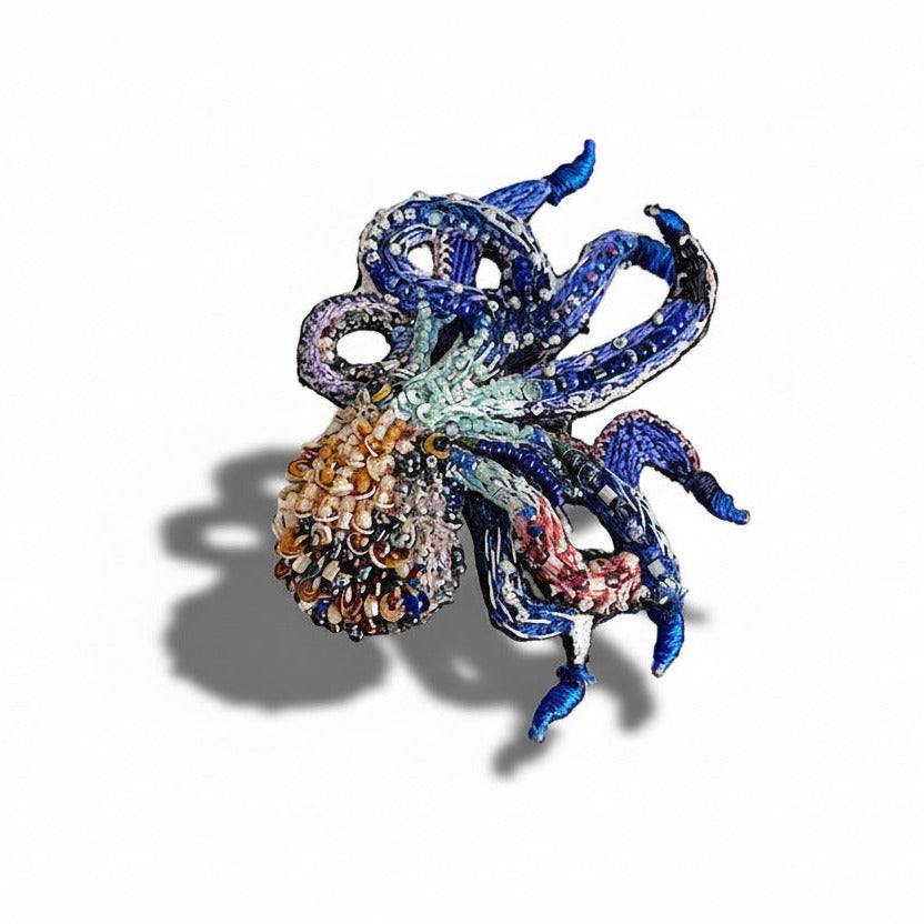 Cette broche sculpturale en textile est ornée de mini perles de verre et de sequins, dans des tons de bleu et de brun. Entièrement réalisée à l’aiguille, ce bijou inspiré par la nature et les fonds marins, est signée Trovelore.