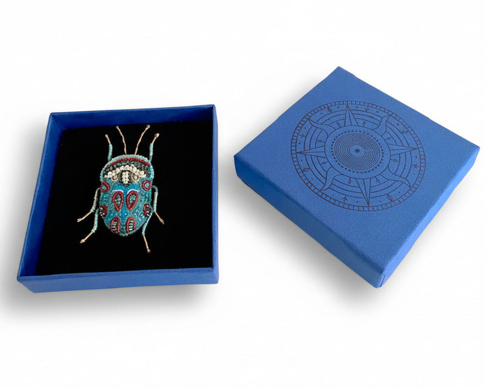 Prête à offrir, La broche Bug Picasso est présentée dans une boîte bleue signé Trovelore, accompagnée de sa notice explicative détaillant le travail de broderie et le savoir-faire artisanal.