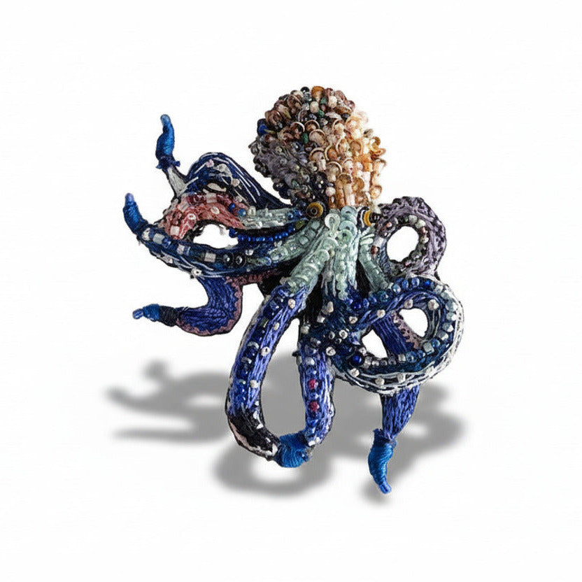 Entièrement réalisée à l’aiguille, cette broche sculpturale en textile est ornée de mini perles de verre et de sequins, dans des tons de bleu et de brun.