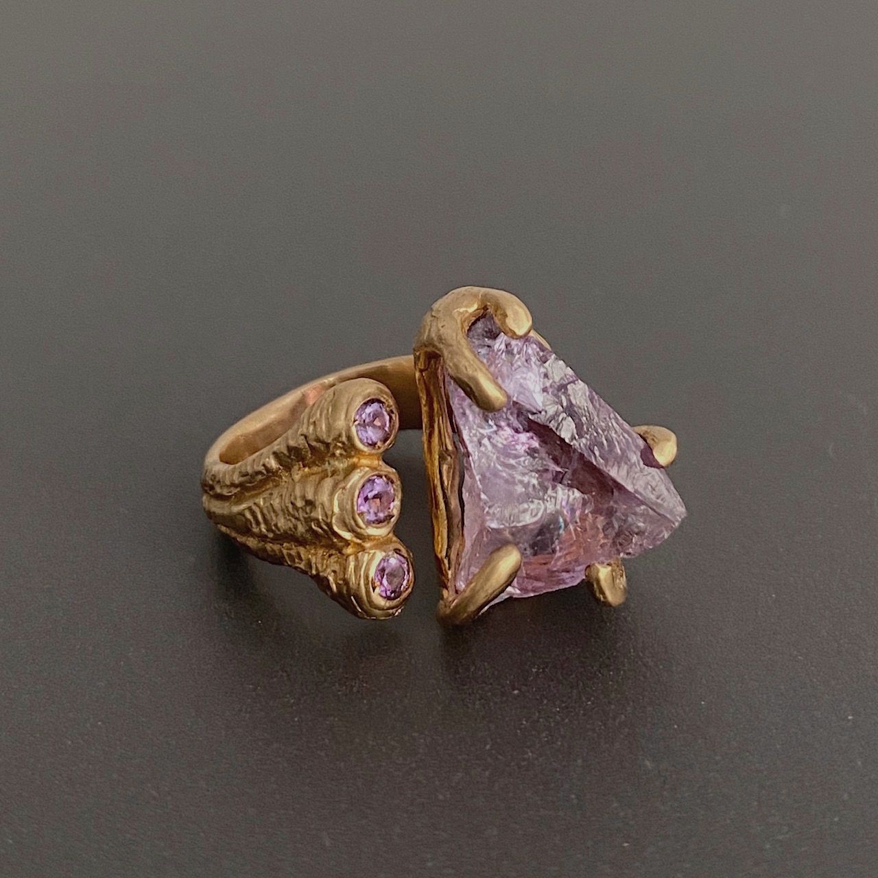 Bague ouverte en bronze sertie d'une imposante améthyste triangulaire brute. A l'autre extrémité de l'anneau trois améthystes taillées contraste le bijou.