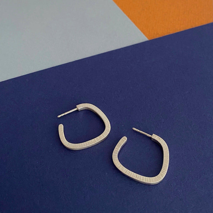 Boucles créoles géométriques en argent forgé et surfaces striées posées sur fond couleurs bleu,gris,orange. Façonnées à la main par la créatrice Anne Milbeau.