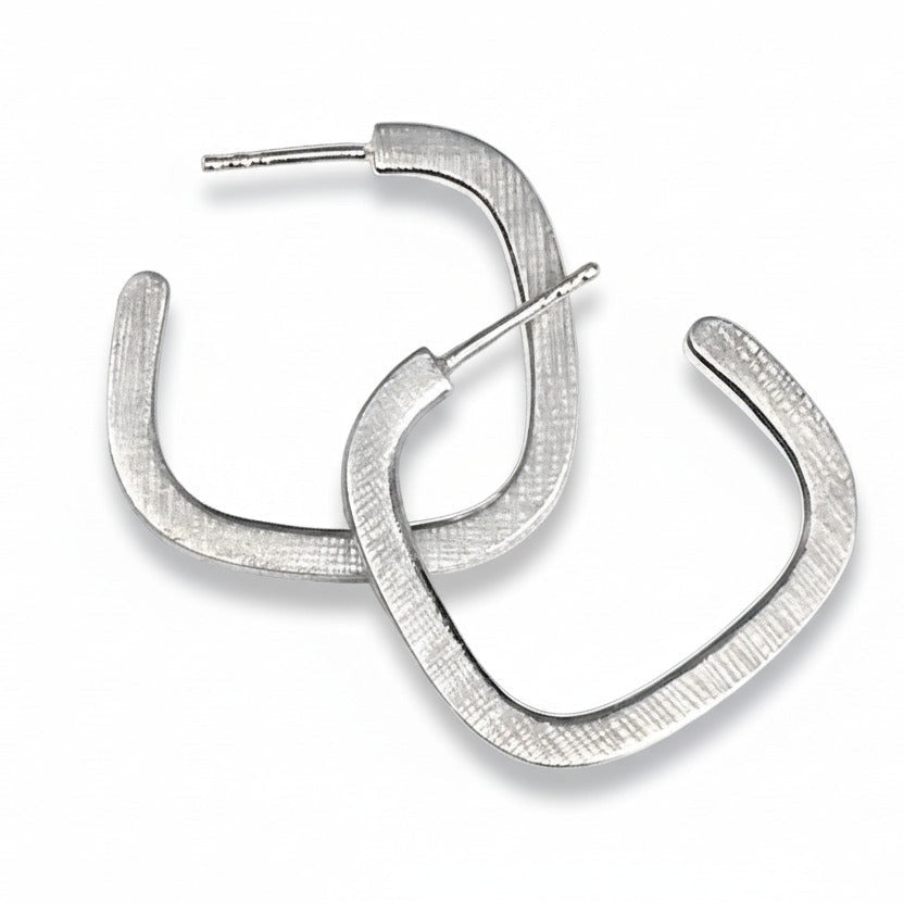 Boucles créoles contemporaines en argent forgé en fil carré. Surfaces striées, intérieur poli : un jeu subtil de lumière et de contraste. Une création minimaliste signée Anne Milbeau.