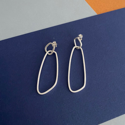 Boucles d’oreilles pendantes en argent satiné de type mobile composées de deux éléments posées géométriques sur fond couleurs bleu, gris et jaune. 