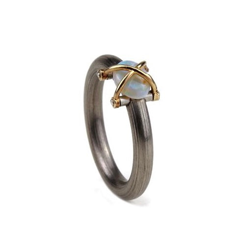 Une Opale en cabochon est présentée sur un anneau en acier inoxydable mat. Deux éléments d'or jaune entrecroisés viennent entrelacer la pierre, comme une cage protectrice. Bijou de profil.